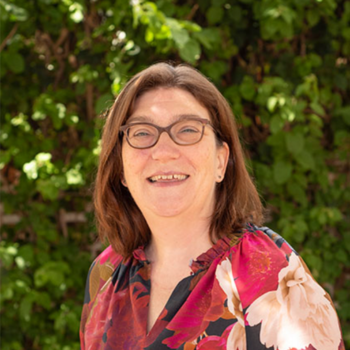 Smiling woman with glasses wearing a floral top, standing outdoors against green foliage.